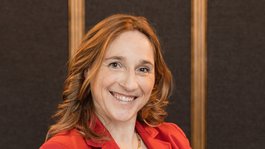 Your hosts in the Dolomites. Hotels + 3 stars? Cristallo! Woman in red blazer smiling with crossed arms against dark background