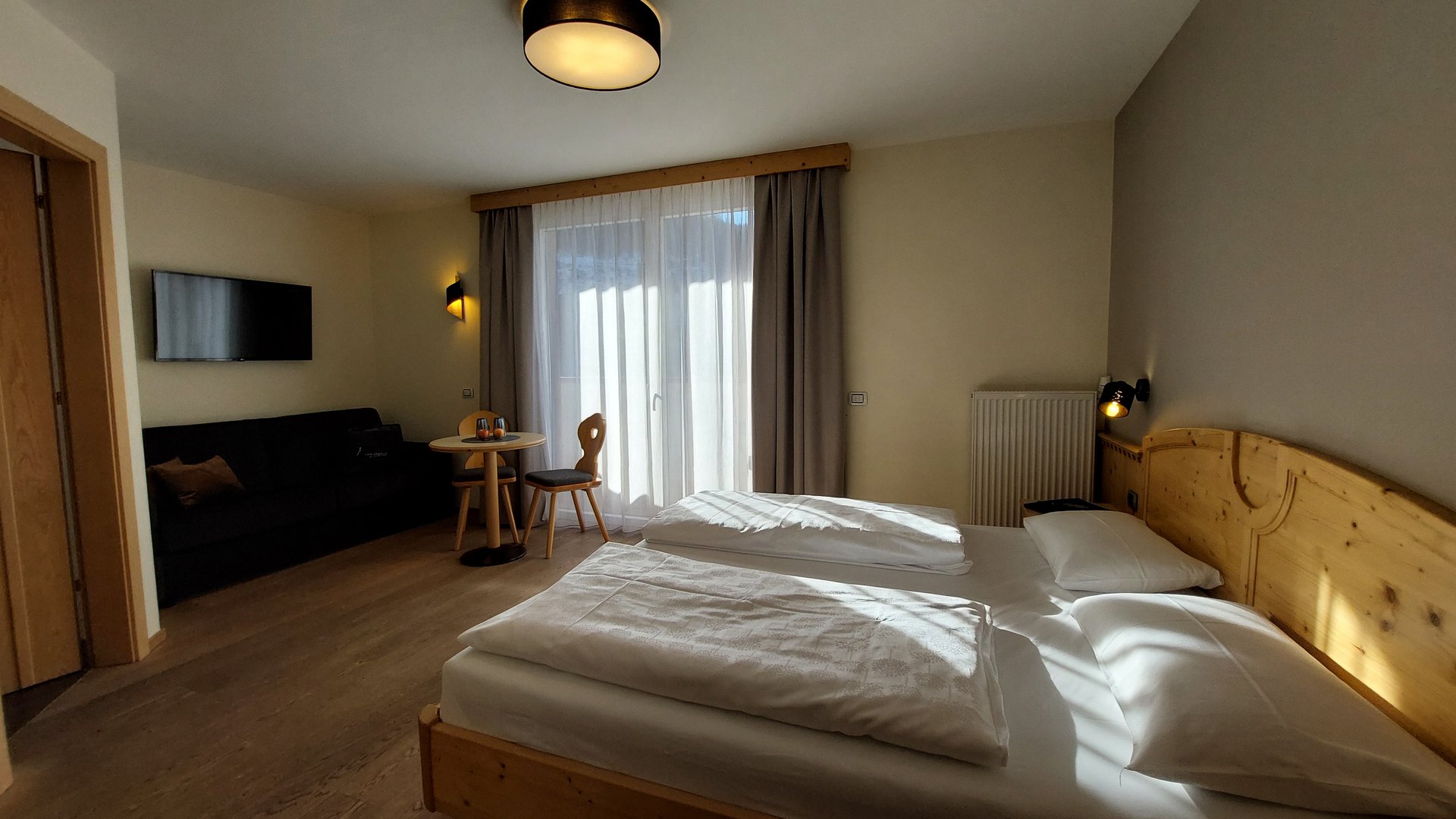 Deine Unterkunft in St. Christina im Grödnertal: Willkommen! Hell eingerichtetes Hotelzimmer mit Doppelbett, Sofa, Tisch und Fernseher