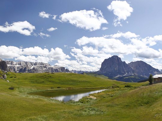 Dolomiten + Wandern + Hotel Cristallo = hallo Sommer! Grüne Wiese mit kleiner Wasserfläche vor Bergkulisse unter blauem Himmel