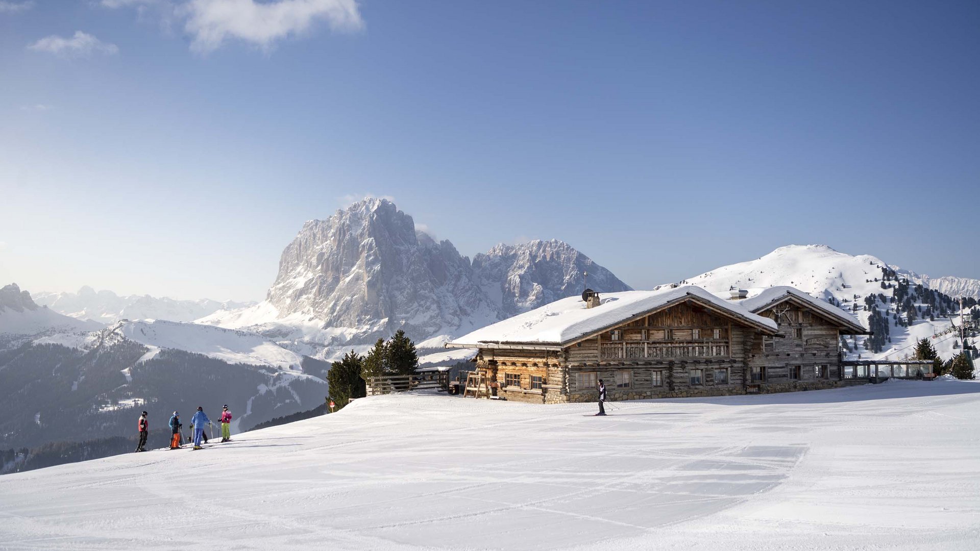 Your hosts in the Dolomites. Hotels + 3 stars? Cristallo! Skiers near a mountain cabin in a snowy landscape under a clear sky