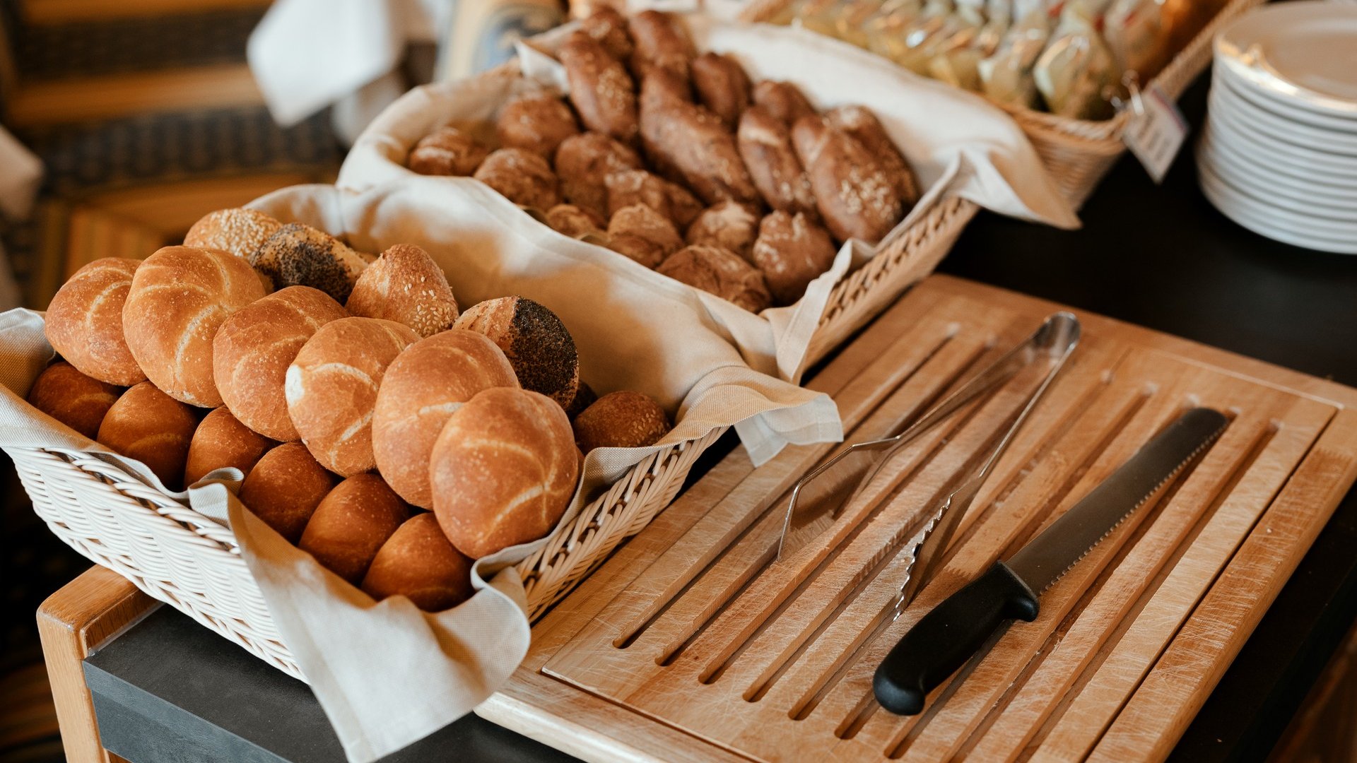 Assorted fresh bread rolls in basket with bread knife and tongs on wooden board