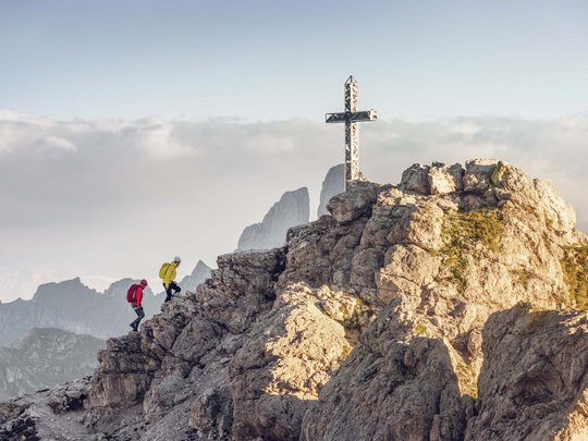 Dolomiten + Wandern + Hotel Cristallo = hallo Sommer! Zwei Wanderer steigen auf felsigen Berg mit Gipfelkreuz bei Bergpanorama