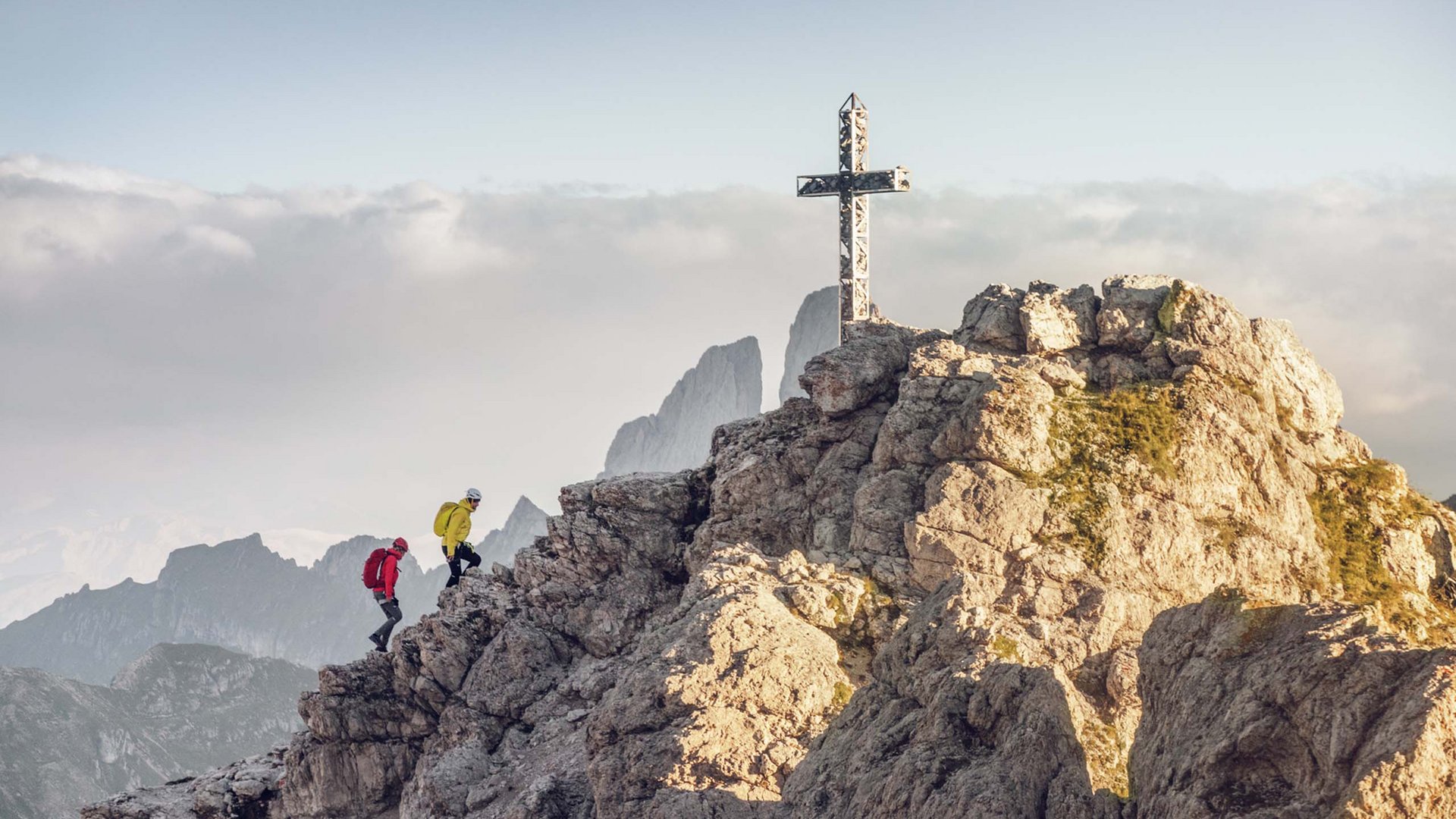 Deine Unterkunft in St. Christina im Grödnertal: Willkommen! Zwei Wanderer steigen auf felsigen Berg mit Gipfelkreuz bei Bergpanorama