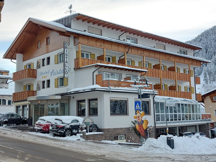 Deine Unterkunft in St. Christina im Grödnertal: Willkommen! Hotel Cristallo im Schnee mit geparkten Autos und verschneiter Landschaft