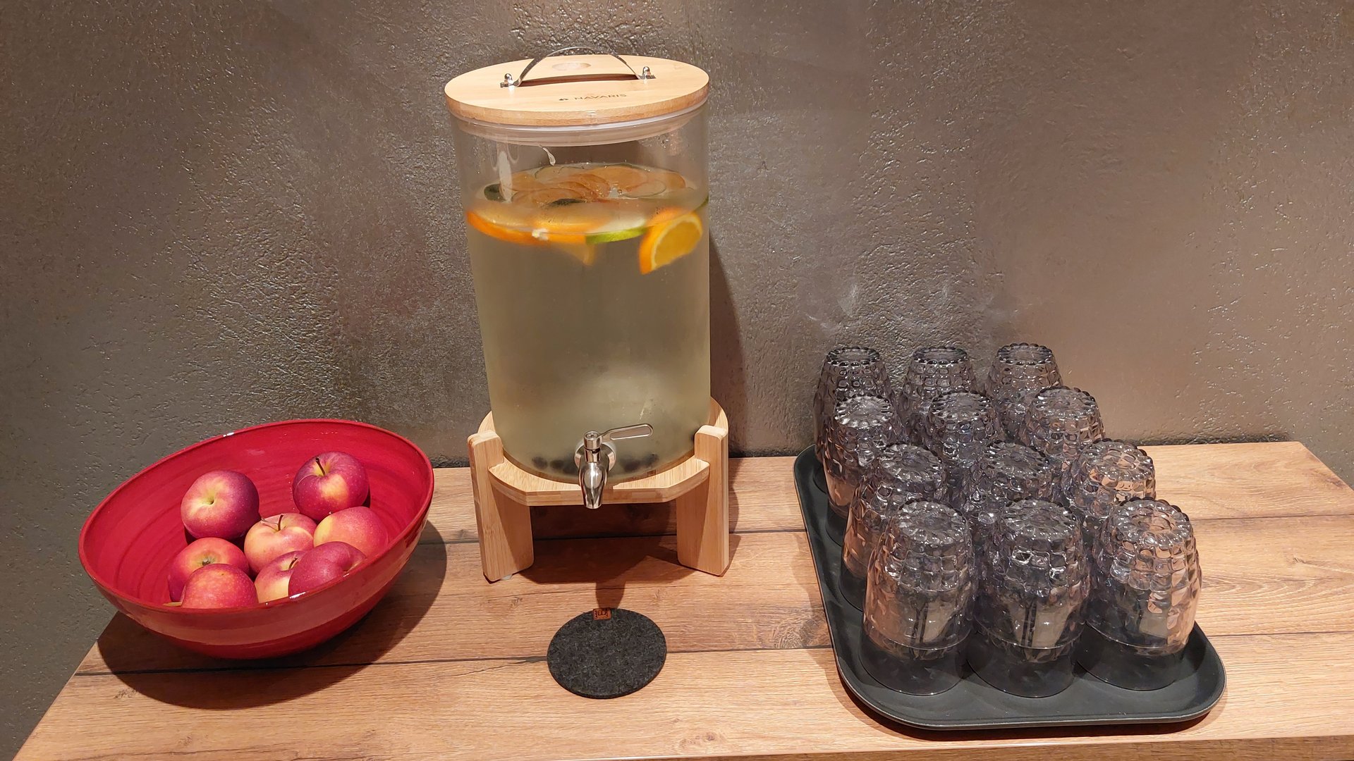 Inklusivleistungen deiner Gröden-Unterkunft Wasserspender mit Früchten, rote Äpfel in Schale und umgedrehte Gläser auf Holzplatte