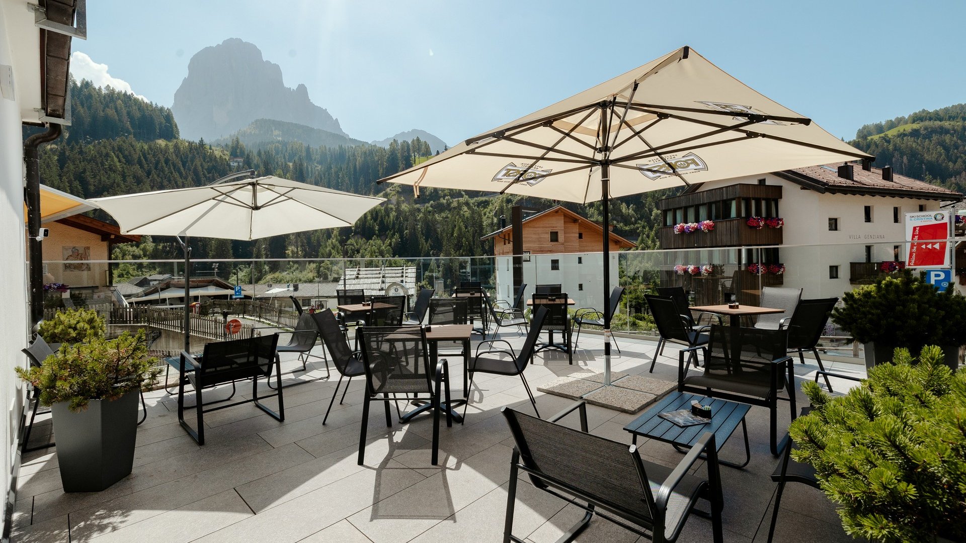 La tua vacanza in Val Gardena è qui Terrazza con tavoli e ombrelloni davanti a paesaggio montano sotto cielo sereno