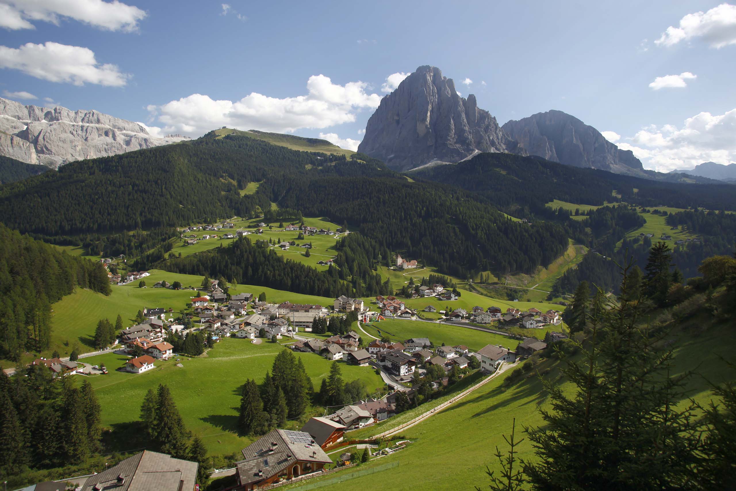 Deine Unterkunft in St. Christina im Grödnertal: Willkommen! Bergdorf mit grünen Wiesen und Felsen in der Ferne unter blauem Himmel